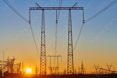 Sun rise on field with high voltage electricity towers. Dark silhouettes of repeating power lines on sunrise. Electricity generation, transmission, and distribution network. Industrial landscape