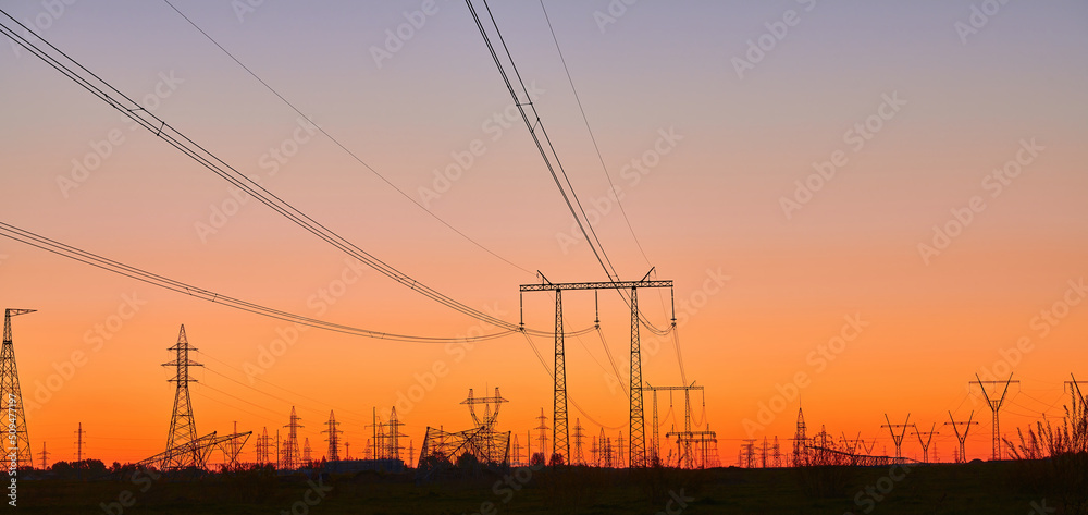 Fototapeta premium High voltage electricity towers on field at sunset and blue sky. Panoramic view of dark silhouettes of power lines on orange sunrise. Electricity generation, transmission, and distribution network