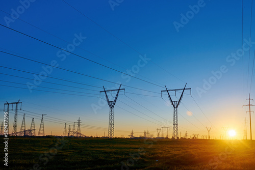 Sun rise on field with high voltage electricity towers. Dark silhouettes of repeating power lines on sunrise. Electricity generation, transmission, and distribution network. Industrial landscape.
