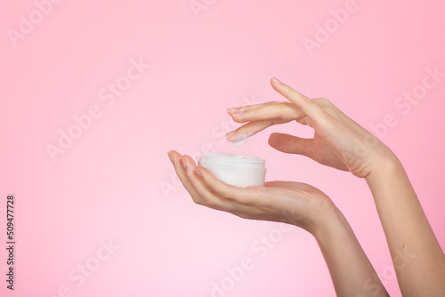 Women's hands with skincare cream jar apply hand care cream, isolated on pink background. Female hands with cosmetic cream. Hydrating and moisture soft face or hand cream . Selective focus, close up