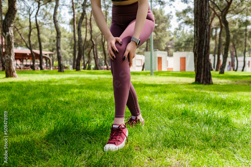 Brunette sportive woman wearing black sports bra standing on city park, outdoors touching injured knee with hands. Injured leg, healthy lifestyle and sport concept.