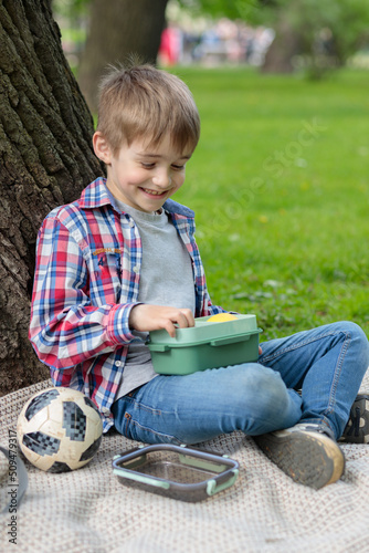 Lunch in the fresh air. A boy sitting by a tree, eating his lunch from a lunch box. Selective focus.