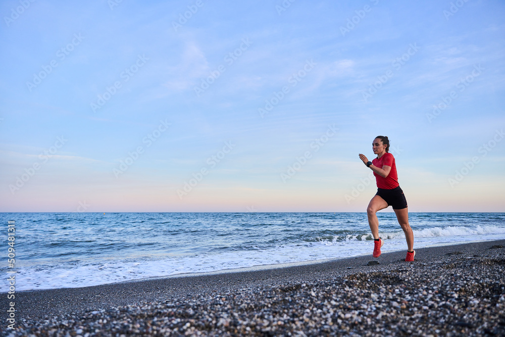 young athletic woman in a red shirt and braid running on the shore of the beach with mountains in the background