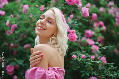 Outdoor portrait of happy smiling beautiful blonde woman wearing pink off shoulder blouse, headband posing in blooming rose garden. Copy, empty space for text 