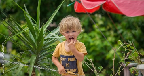 little blond baby boy points his finger to his mouth in summer garden