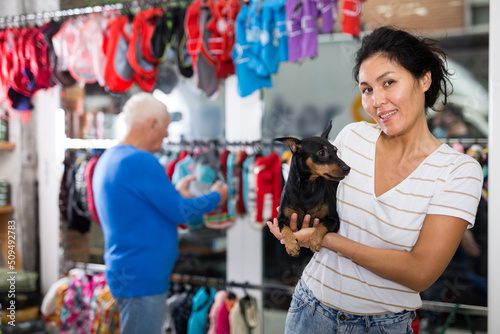 Asian woman with miniature pinscher in hands standing in salesroom of pet shop. Elderly man making purchases in background.