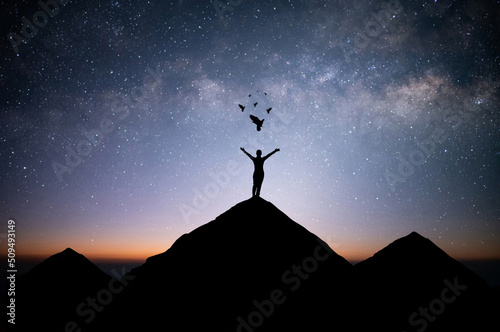 Young woman traveler standing alone on top of mountain and raise both arms praying and free bird enjoying nature on beautiful night sky, star, milky way background. Demonstrates hope and freedom.