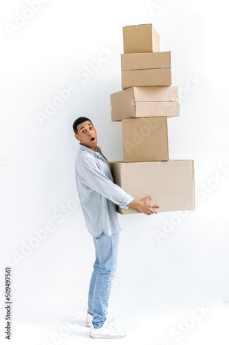 Stunned mixed race guy in casual stylish clothes, delivery man, standing over isolated white background, holding many cardboard boxes, delivering parcels, looking in amazement at camera, delivery