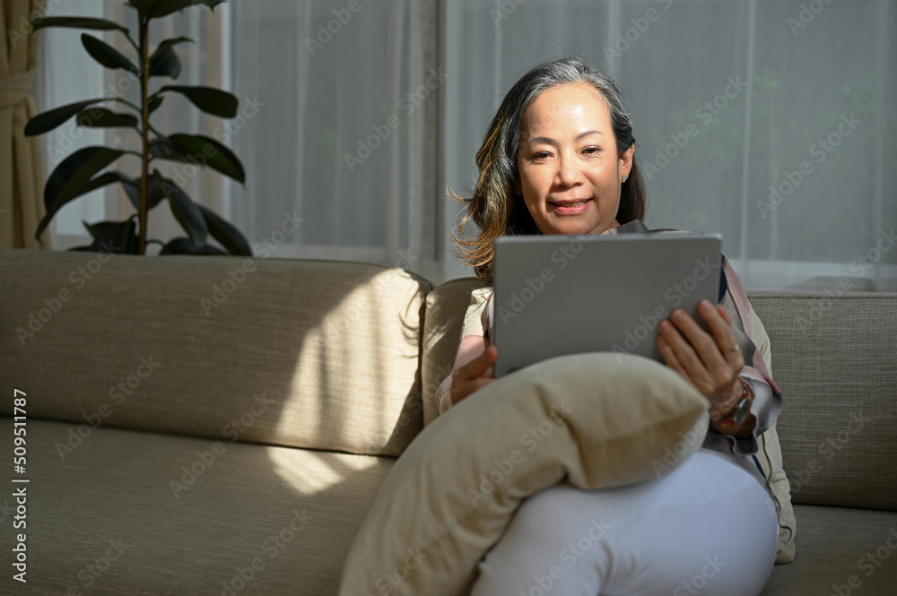© bongkarn - Relaxed asian aged-woman resting in her living room, sitting on comfy sofa and using tablet