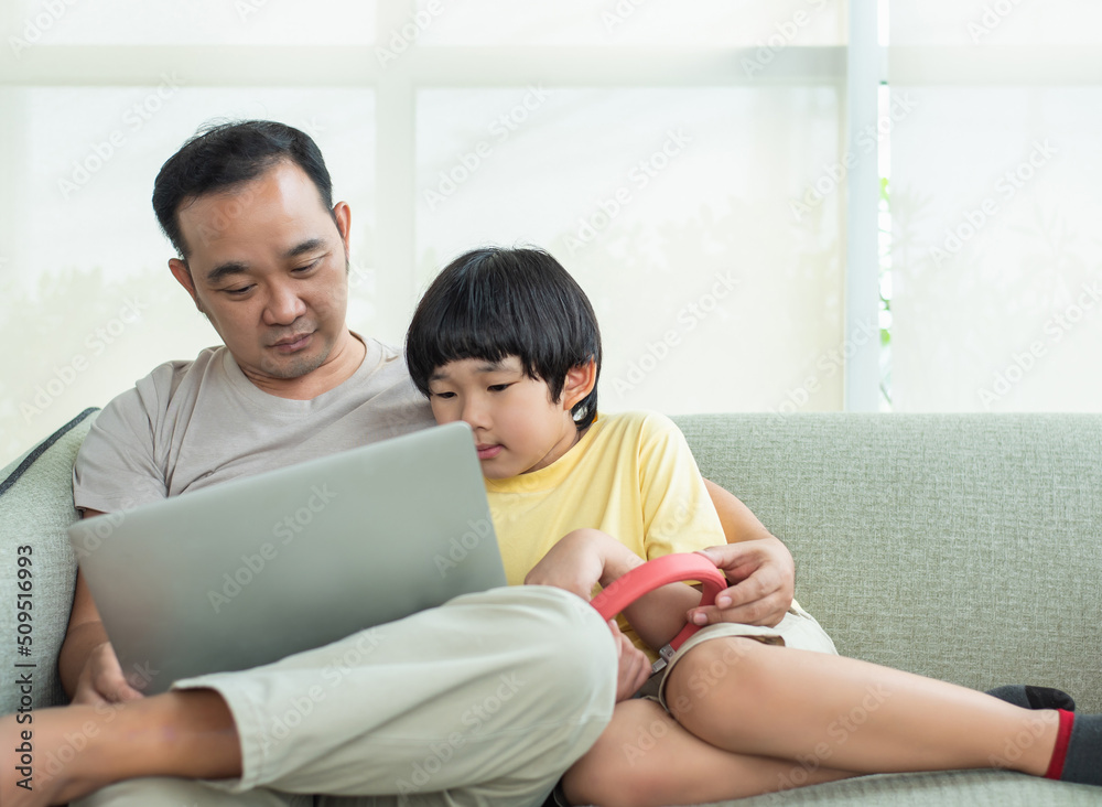 Little boy relaxing on the sofa at home with his father watching funny videos on laptop together. A father is so happy with his preschool son in the living room. Little boy relaxing on the sofa at home with his father watching funny videos on laptop together. A father is so happy with his preschool son in the living room.