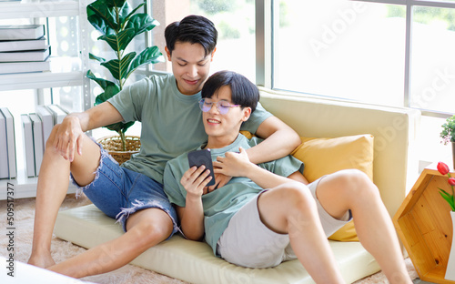 Two Asian young handsome pride male gay men lover couple partner sitting smiling laying down together on cozy sofa in bedroom cuddling hugging watching entertainment media streaming on smartphone © Bangkok Click Studio