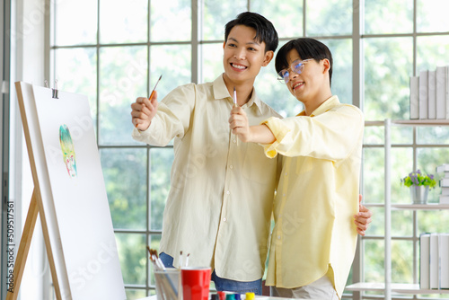 Two Asian young lovely pride teenager male gay men lover couple partner standing smiling having fun playing paintbrush in front canvas painting board on easel in glass windows workshop studio at home