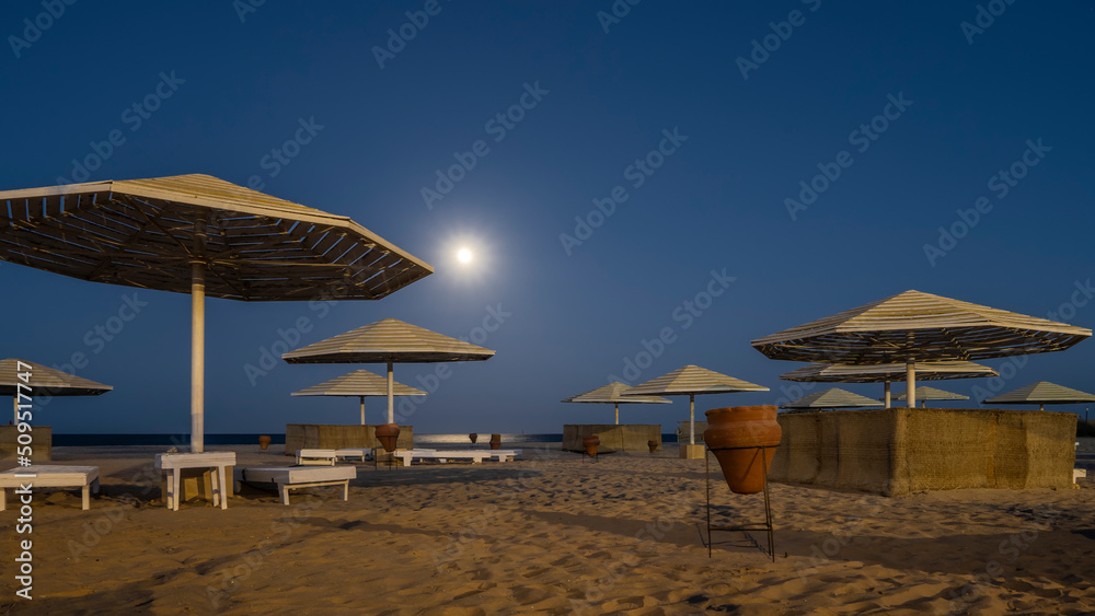 Fototapeta premium A quiet night on the Red Sea beach. The full moon is shining in the dark blue sky. Lattice umbrellas, empty sunbeds, urns are on the sand. Egypt. Safaga