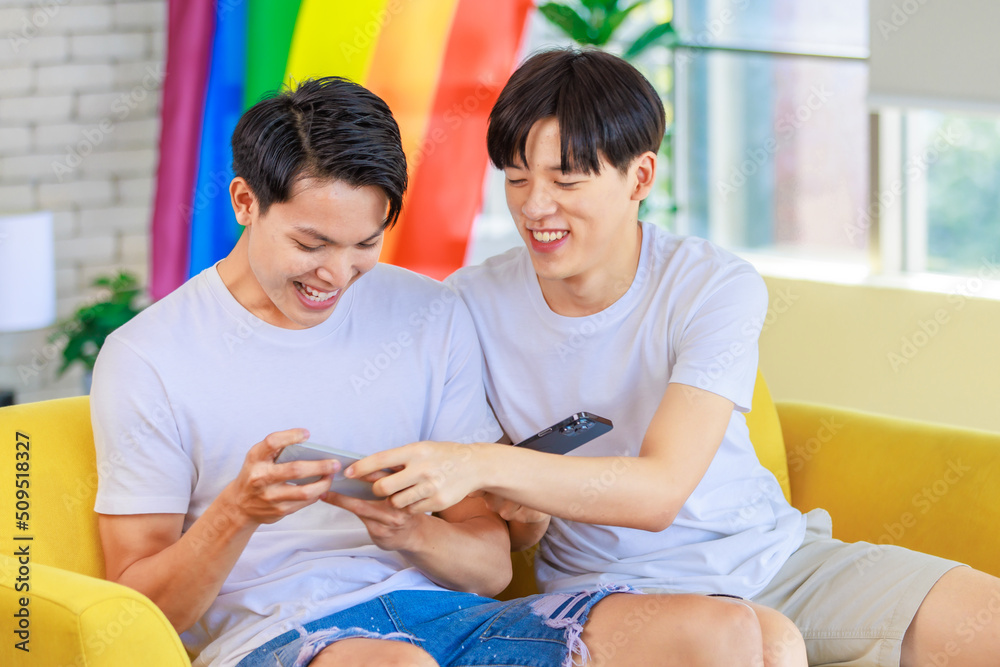 Two Asian cheerful teenager male gay men lover couple partner sitting smiling on sofa holding smartphone playing fun digital game online together in living room with rainbow pride equality unity flag