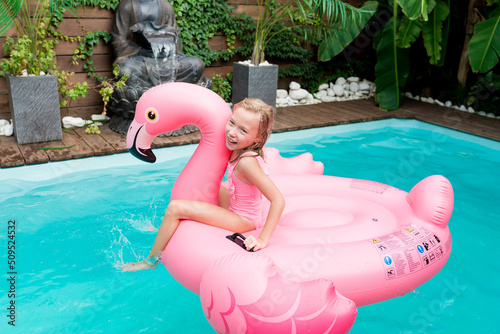 Outside leisure and happy childhood concept. Cute child girl sitting on inflatable pink flamingo while swimming in the pool on summer day.