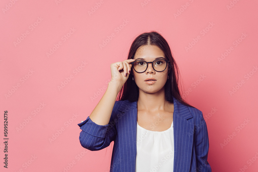 Young brunette in glasses dressed in a dark blue jacket and white blouse, adjusts her glasses, looking seriously, over pink background with copy space. Smart people concept. Young brunette in glasses dressed in a dark blue jacket and white blouse, adjusts her glasses, looking seriously, over pink background with copy space. Smart people concept.