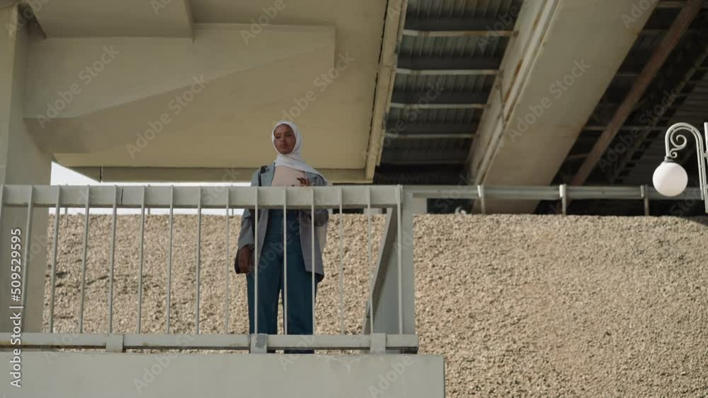 Happy Muslim lady in casual clothes waves hand greeting friend standing on ground under bridge. African-American woman in modern city. Travelling and vacation Happy Muslim lady in casual clothes waves hand greeting friend standing on ground under bridge. African-American woman in modern city. Travelling and vacation