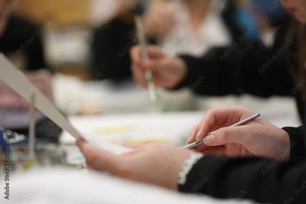 A close up of an art students hand holding a small grey water color paint brush painting at a desk in a school art class room Other students painting deliberately blurred in the background.