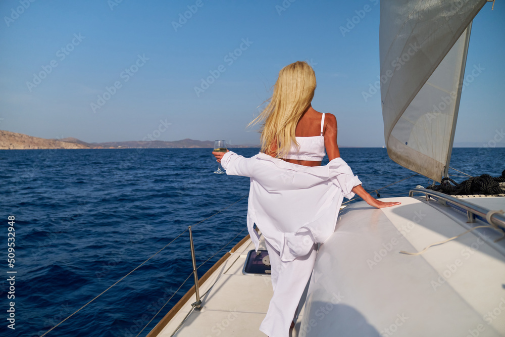 Obraz premium Beautiful young blond woman standing on catamaran at sunny summer day and holding glass of wine