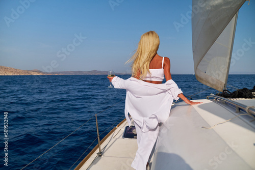Beautiful young blond woman standing on catamaran at sunny summer day and holding glass of wine © Anatoly Repin