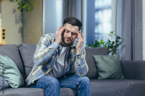 The sick man is sitting alone at home on the couch, has a severe headache