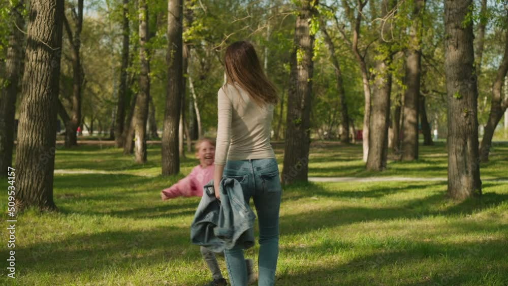 Little girl with long plait runs around happy mother joining hands in spring park slow motion. Woman with brownish hair plays with blonde daughter in garden Little girl with long plait runs around happy mother joining hands in spring park slow motion. Woman with brownish hair plays with blonde daughter in garden