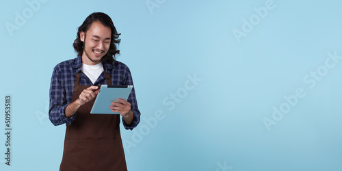 Obraz na plátne Young asian entrepreneur man in apron hand holding tablet for list order food isolated on blue background