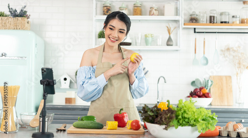Asian woman food blogger cooking salad in front of smartphone camera while recording vlog video and live streaming at home in kitchen. photo