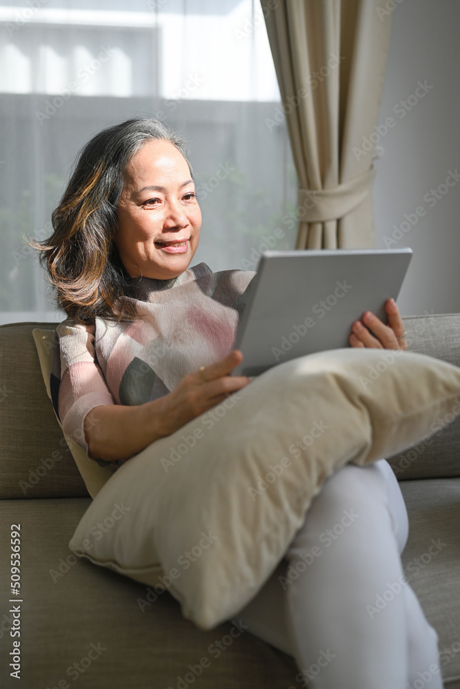 Positive middle aged woman browsing wireless Internet on tablet, relax on sofa at home. Elderly technology concept Positive middle aged woman browsing wireless Internet on tablet, relax on sofa at home. Elderly technology concept