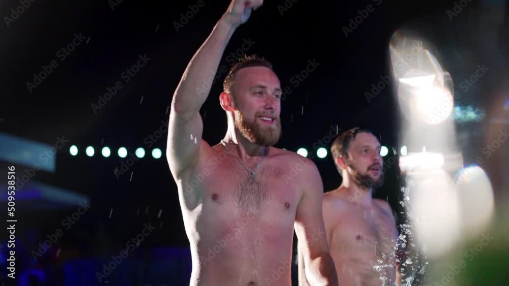 Happy homosexual man celebrating gay pride with friends at night LGBTQ pool party in a private villa swimming pool. Bisexual male in swimwear dancing splashing water in luxury resort. Slow motion.