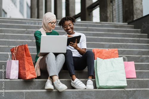Fotografering Two pretty multiracial young best friends women, making online purchases using a tablet and laptop sitting on the steps of the mall with colorful shopping bags, having fun and laughing together
