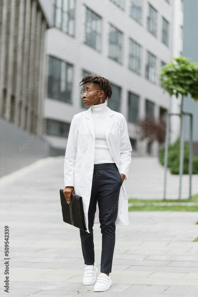 Portrait of young smart focused african female medical intern doctor with folder clipboard, standing outside modern hospital building. Medicine, education and healthcare concept.