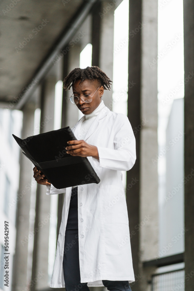 Young handsome skilled female African heathcare worker, making notes on folder clipboard, standing on stairs of modern building. Professional doctor filling in medical record outdoors Young handsome skilled female African heathcare worker, making notes on folder clipboard, standing on stairs of modern building. Professional doctor filling in medical record outdoors