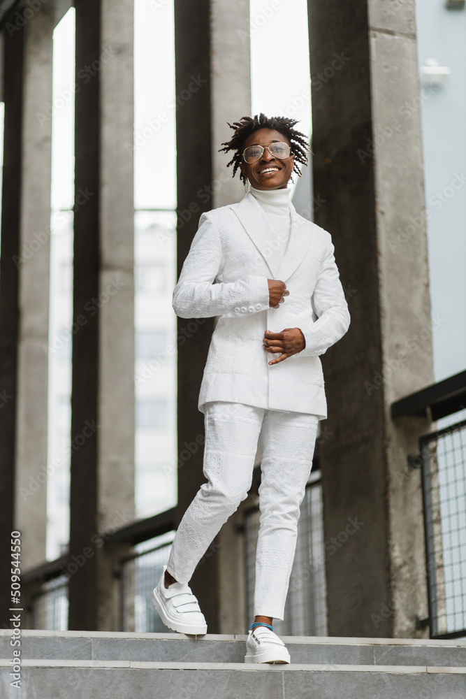 Portrait of smiling joyful beautiful fashionable African woman, wearing eyeglasses, white stylish suit, standing on modern city street. New modern buildings, blurred background with columns. Portrait of smiling joyful beautiful fashionable African woman, wearing eyeglasses, white stylish suit, standing on modern city street. New modern buildings, blurred background with columns.