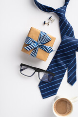 Father's Day concept. Top view vertical photo of craft paper giftbox with ribbon bow blue tie glasses cup of coffee and cufflinks on isolated white background
