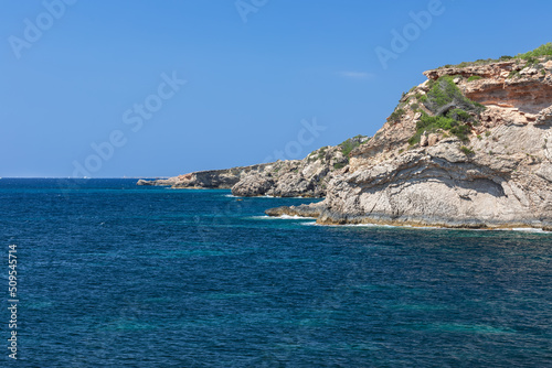 Rocky coast of Ibiza island scorched by the summer sun with sparse vegetation, calm peaceful sea, clear day, not a cloud in the sky, Balearic Islands, Spain