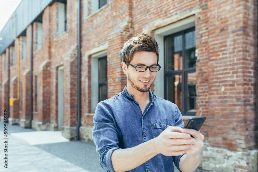 Fototapeta premium Portrait of young male student, outside campus holding cellphone, smiling and walking