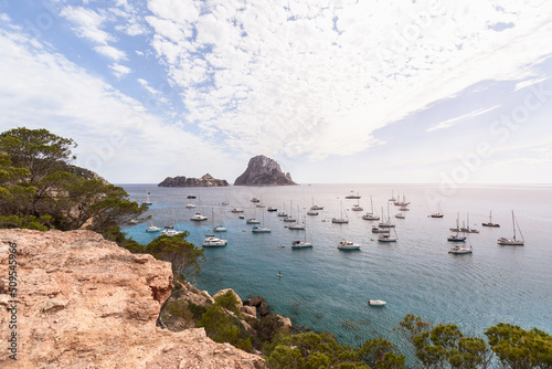 Panoramic shot of coast dropping into waters of bay Cola d'Hort and numerous anchored sailing yachts and catamarans. Ibiza, Balearic Islands, Spain