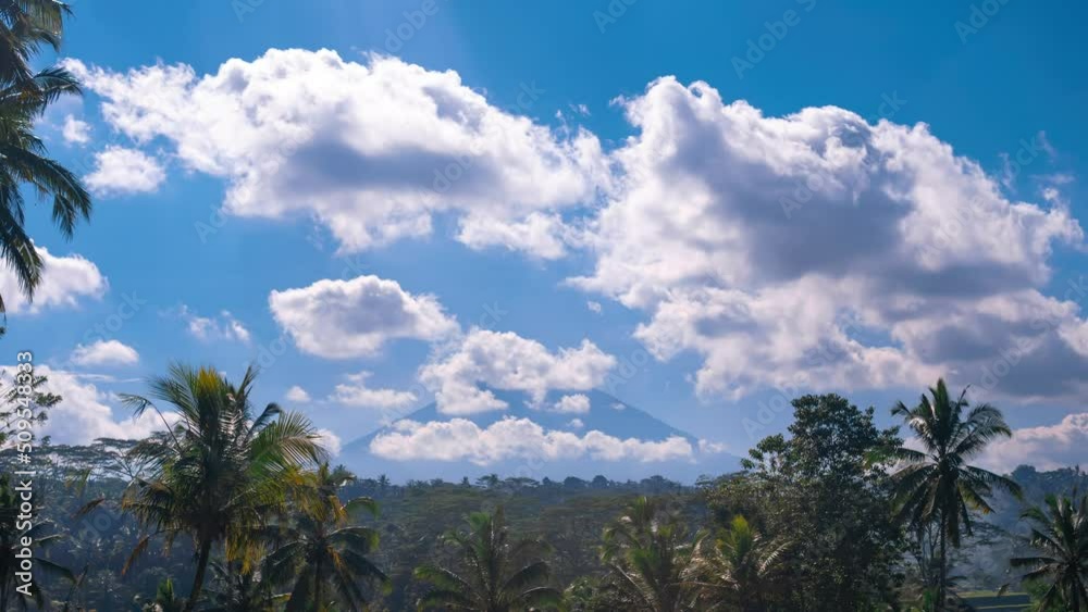 Timelapse of clouds moving from wind in blue sky on sunny day with a volcano on background. Palm trees, tropical area