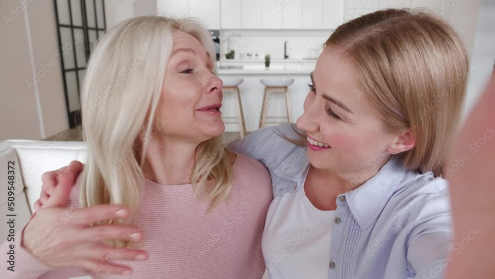 Happy affectionate mature mother and grownup daughter make selfie video call by mobile phone while sitting on cosy sofa at home. Happy affectionate mature mother and grownup daughter make selfie video call by mobile phone while sitting on cosy sofa at home.