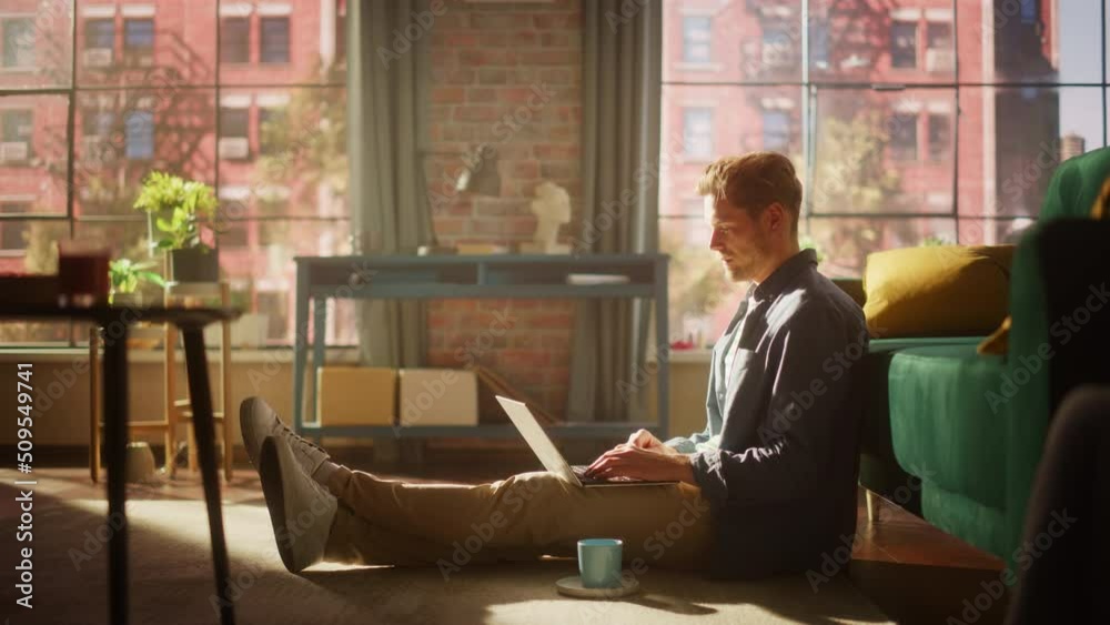 Handsome Young Man Using Laptop, Working from Home Living Room with Big Windows. Male Sitting on Floor, Using Computer, Writing Project Plan. Successful Manager Doing Remote Work. Handsome Young Man Using Laptop, Working from Home Living Room with Big Windows. Male Sitting on Floor, Using Computer, Writing Project Plan. Successful Manager Doing Remote Work.
