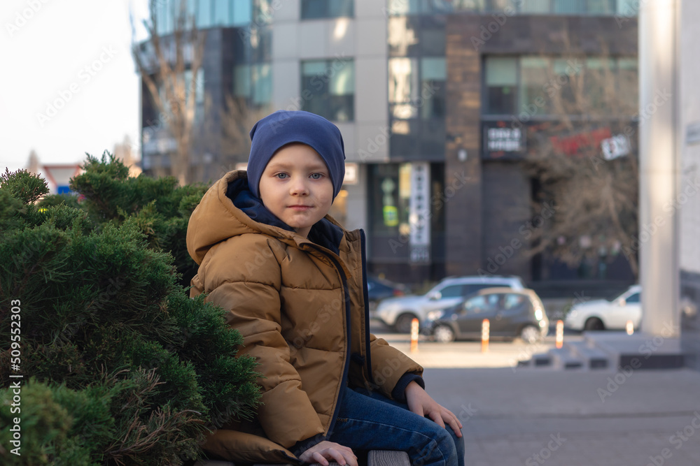 Fototapeta premium a boy in a jacket and hat sits on a bench in the city