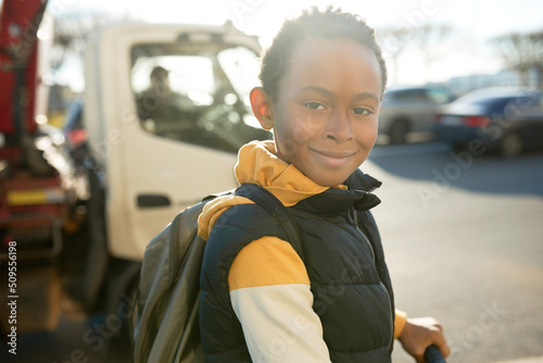 Outdoor portrait of cute active african american boy in yellow stylish hoodie and black puffer vest riding scooter, standing over city background, waiting to cross road on traffic jam