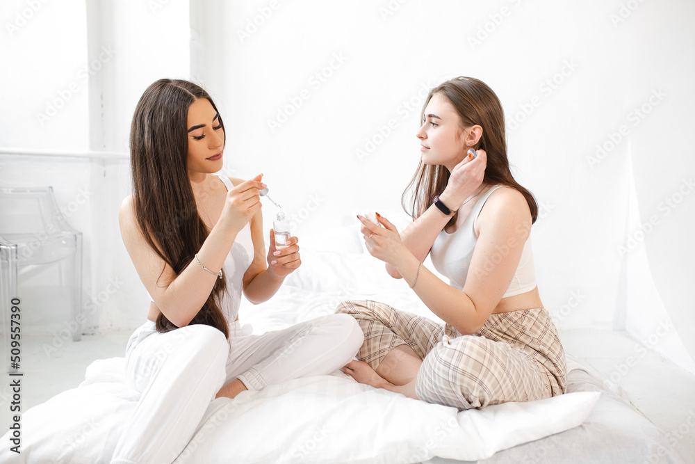 Two young beautiful girls with cosmetics. Women make face masks to take care of their skin. Girlfriends in a home spa rest and relax on a white bed