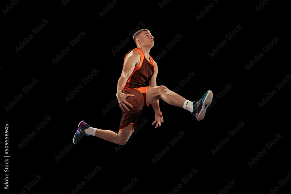 Dynamic portrait of young professional basketball player in motion, jumping with a ball isolated over black studio background