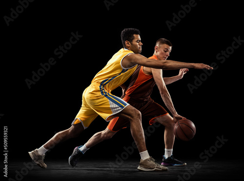 Portrait of two sportive men, basketball players in uniform playing, training isolated on black studio background. Following the ball carrier.