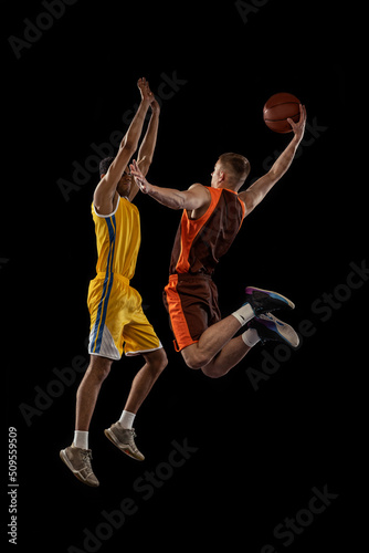 Portrait of two young men, professional basketball players in a jump, throwing ball into basket isolated over black studio background. Winner