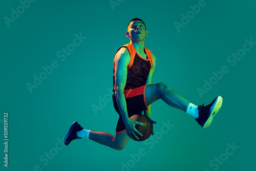 Portrait of young man, professional backetball player in motion, training isolated over blue background in neon light. Jump shot