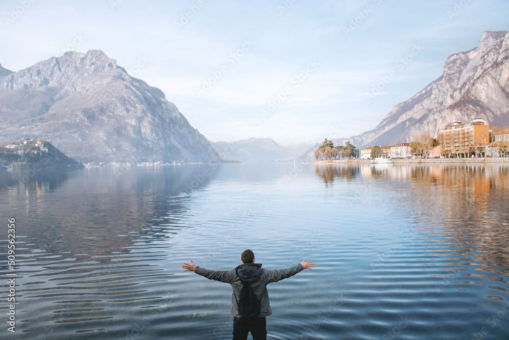 Blurred focus. Man traveler enjoys freedom with raised hands and backpack on the shores of Lake Lecco, Italy. Blue water, mountains and sky. Concept of tourist lifestyle, summer vacation outdoors.