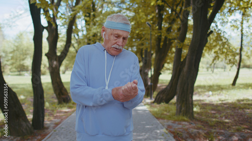 Fototapeta Naklejka Na Ścianę i Meble -  Cinematic footage of motivated athletic fitness retired senior mature male jogger in sportswear running checking pulse on arm in park. Active elderly grandfather man enjoying healthy sport lifestyle
